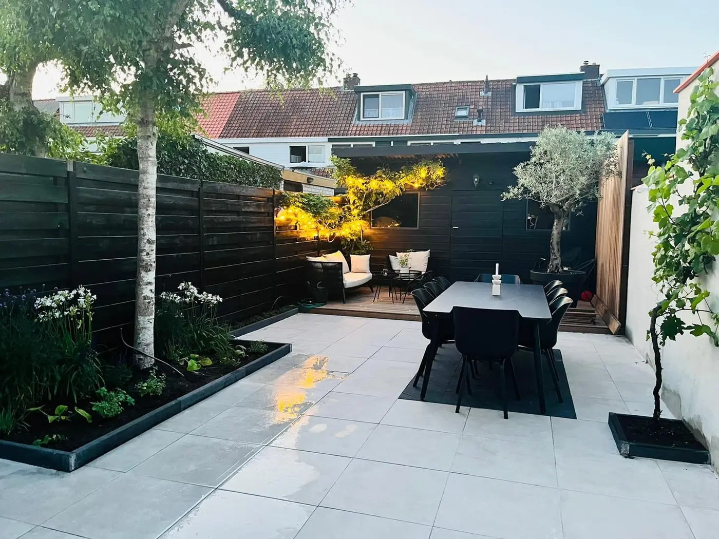 Dezelfde achtertuin na de renovatie: zwarte betimmering, keramische tegels, olijfboom en lichtsnoeren